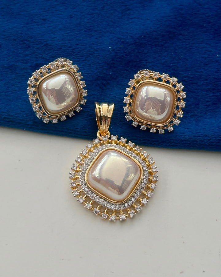 A gold-plated jewelry set featuring a pendant and matching earrings, each with a square iridescent pearl surrounded by small clear stones, displayed on a blue velvet surface.