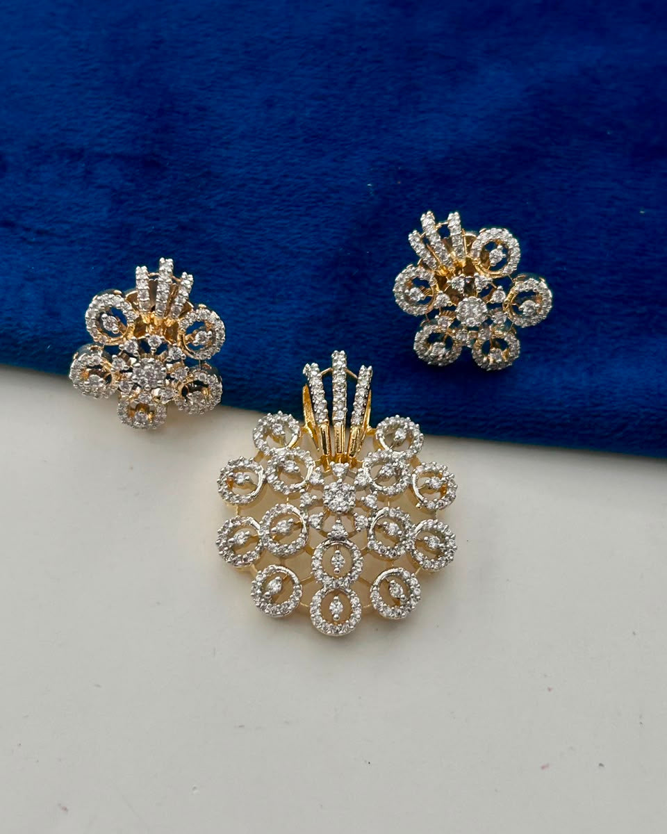 A gold and silver-tone jewelry set featuring a large floral cluster pendant and two matching stud earrings, decorated with clear stones, displayed on a split white and dark blue velvet background.