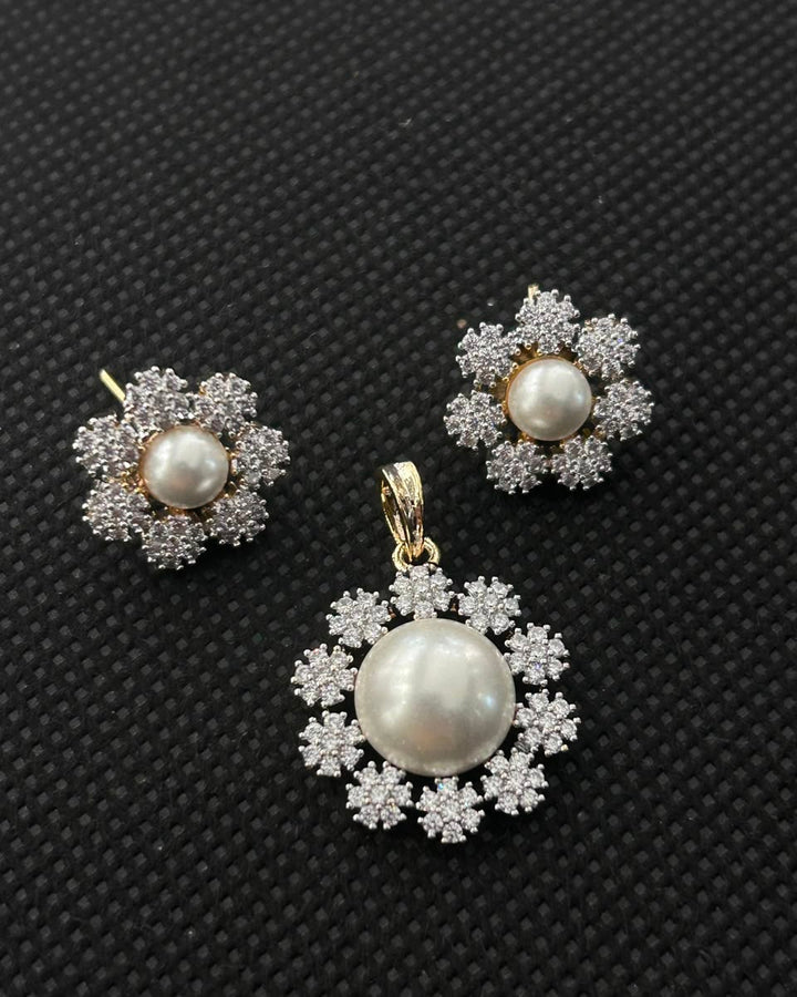 A jewelry set featuring a pendant and matching stud earrings, each with a central white pearl surrounded by a halo of small clear stones in a gold-tone setting, displayed on a textured black background.