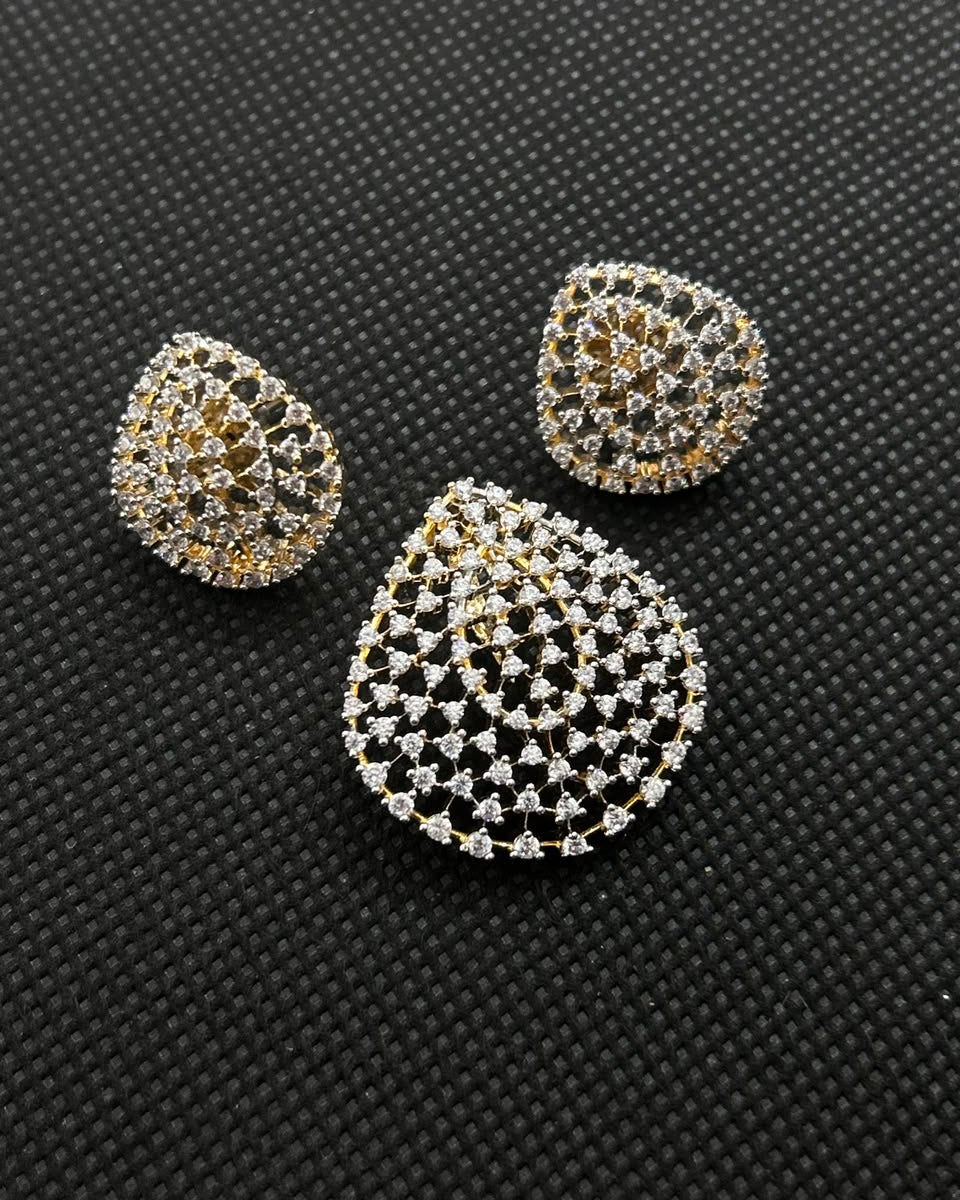 A jewelry set featuring a large teardrop-shaped brooch and two matching teardrop-shaped earrings, all adorned with intricate silver and gold filigree patterns set with clear stones, displayed on a textured black background.