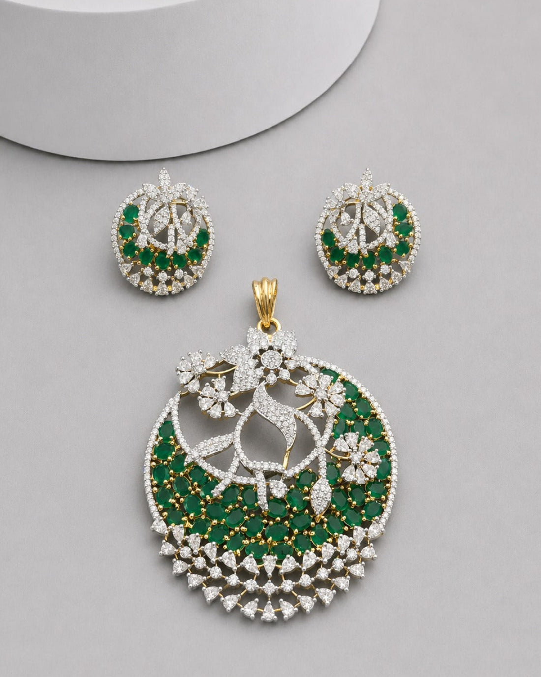 A gold and green gemstone jewelry set featuring a large circular pendant, a matching pair of earrings, and a small box, displayed on a light gray surface with a white cylindrical prop.