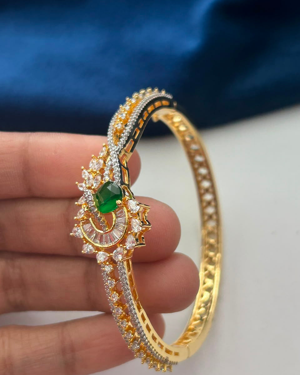 A gold-plated bangle bracelet with intricate crystal and green stone detailing, held by a hand against a dark blue fabric background.