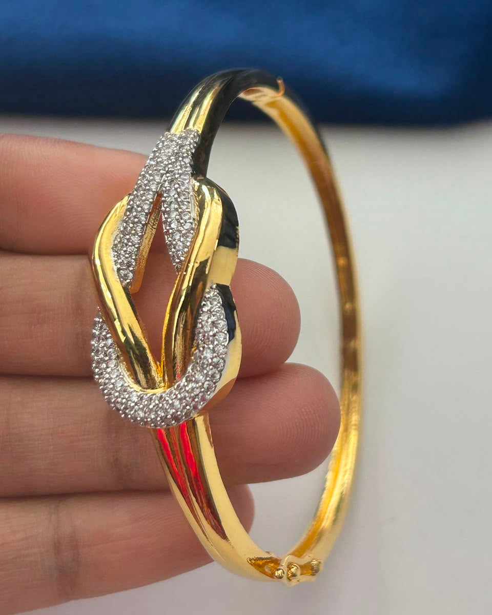 A gold hinged bangle bracelet with a knot-like central design, featuring pavé-set clear stones on one side and a polished gold finish on the other, held in a hand against a white and blue background.