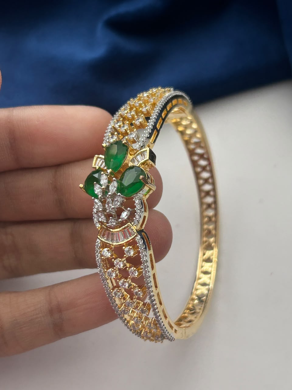 A gold-plated bangle bracelet with green emerald-cut gemstones and clear pavé accents, held by a hand against a white and blue background.