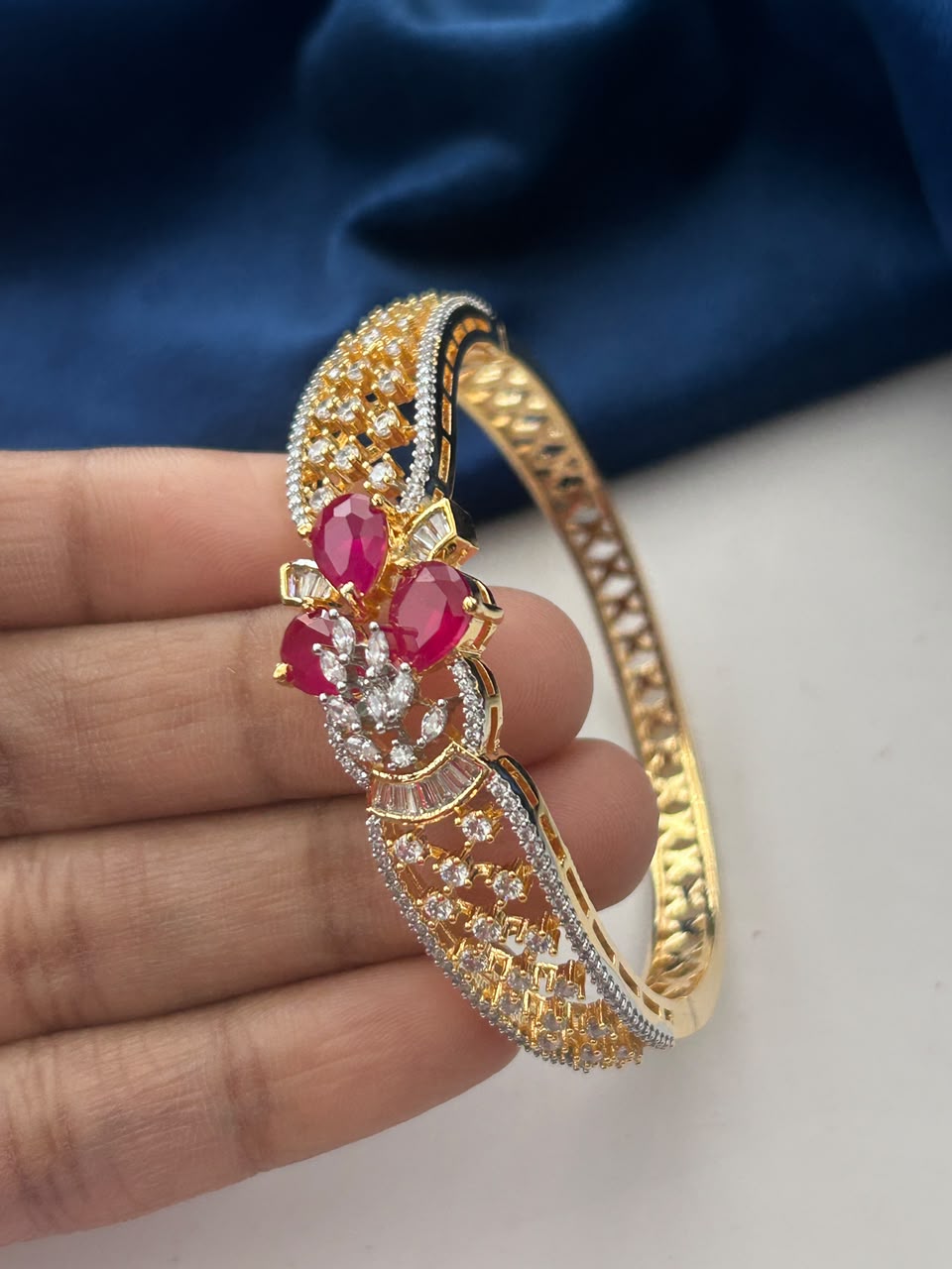 A gold-plated bangle bracelet with intricate pavé detailing, featuring three prominent red gemstone floral motifs, held by a hand against a dark blue fabric background.