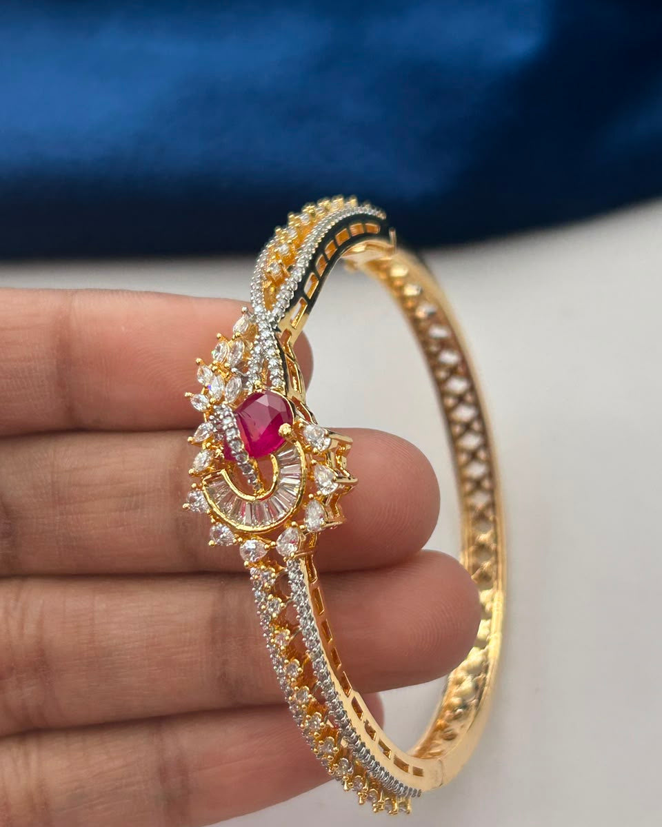A gold-plated bangle bracelet with a central red gemstone and clear stones, held in a hand against a blue and white background.