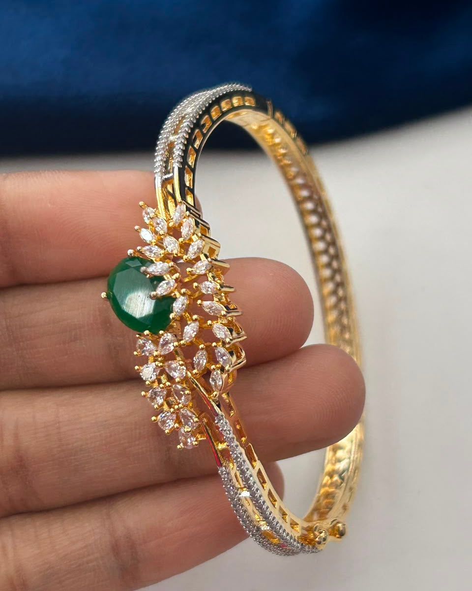 A close-up of a gold-plated bangle bracelet with a green cabochon stone and clear pavé accents, held in a person's hand against a dark blue fabric background.