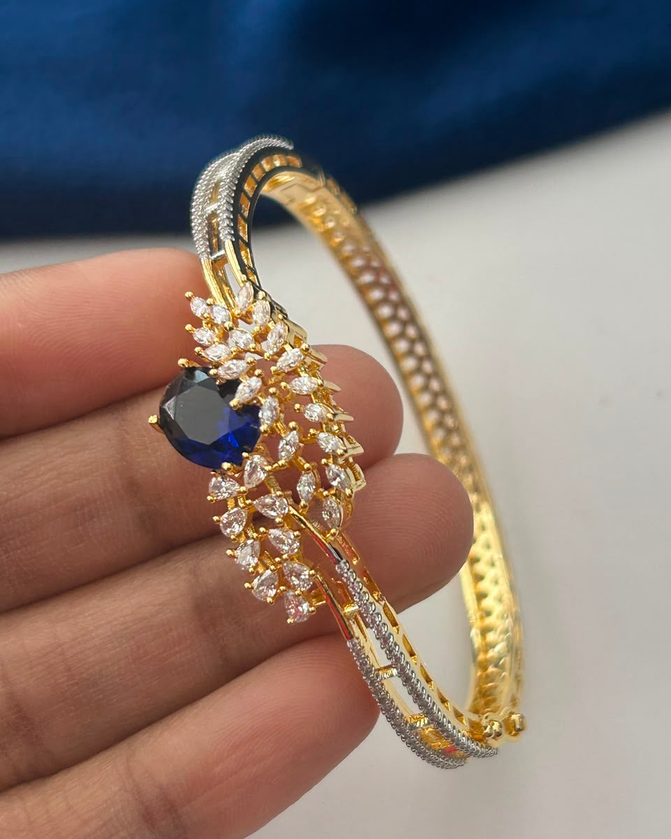 A gold-plated bangle bracelet with a central floral cluster of clear stones and a dark blue oval gemstone, held in a person's hand against a white and blue background.