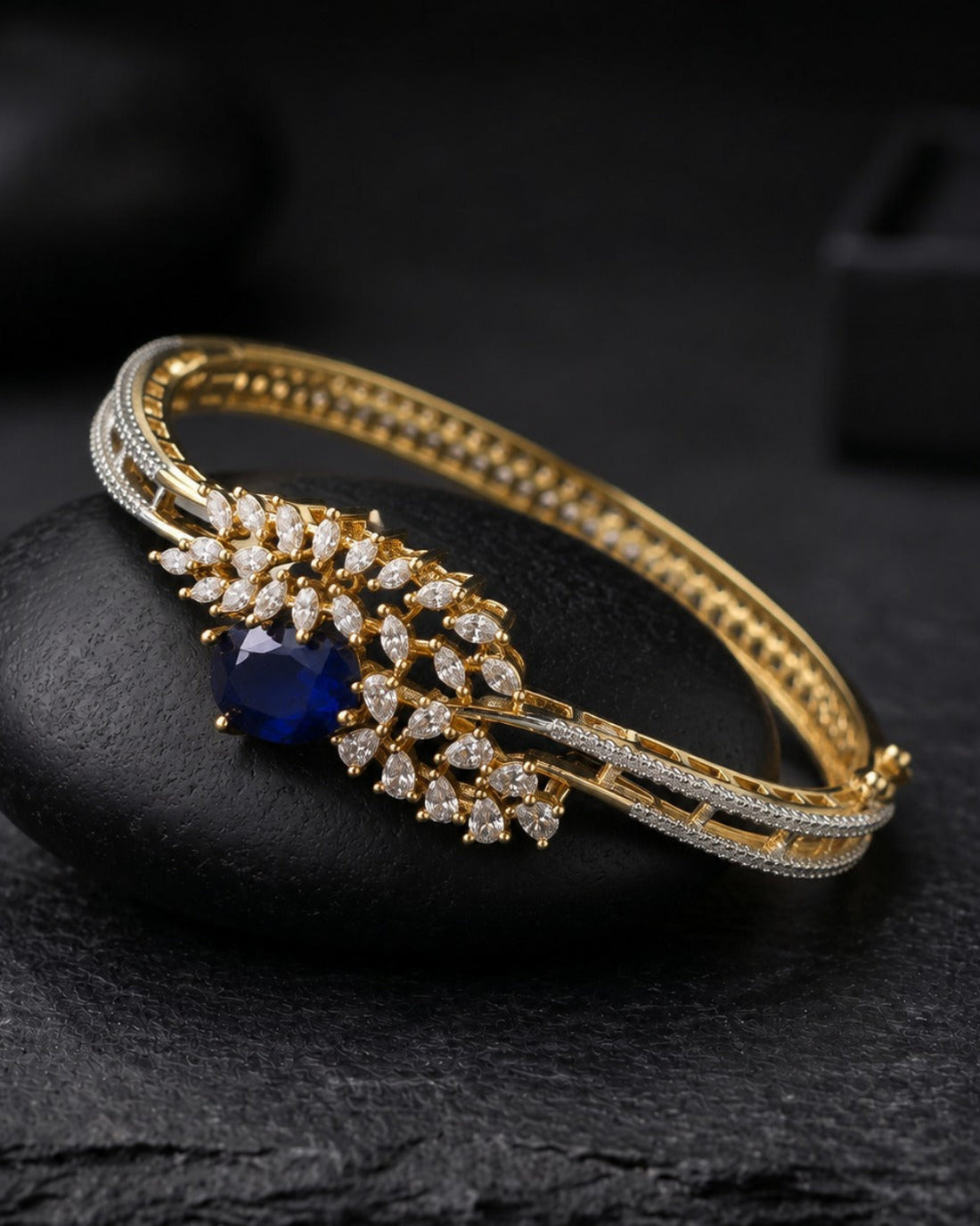 A gold-plated hinged bangle bracelet with a central leaf-shaped cluster of clear pavé stones and a deep blue oval center stone, displayed on a black stone against a dark background.