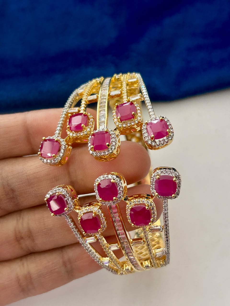 A pair of gold-plated multi-band bangles decorated with square pinkish-red gemstones and small clear stones, held in a person's hand against a blue and white background.