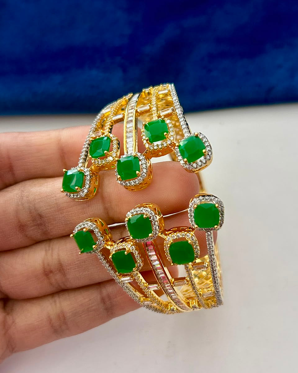 A gold-plated multi-band cuff bracelet with green gemstone accents and small clear stones, held in a hand against a blue and white background.