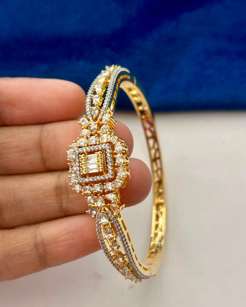 A gold-plated hinged bangle bracelet with intricate diamond and crystal detailing, held in a person's hand against a blue and white background.