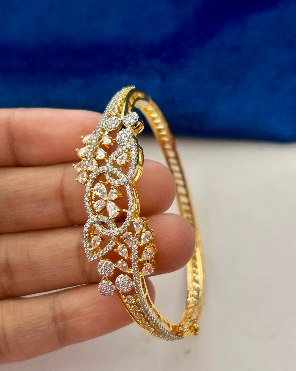 A gold-plated hinged bangle bracelet decorated with intricate pavé diamond floral motifs, held in a hand against a blue velvet background.