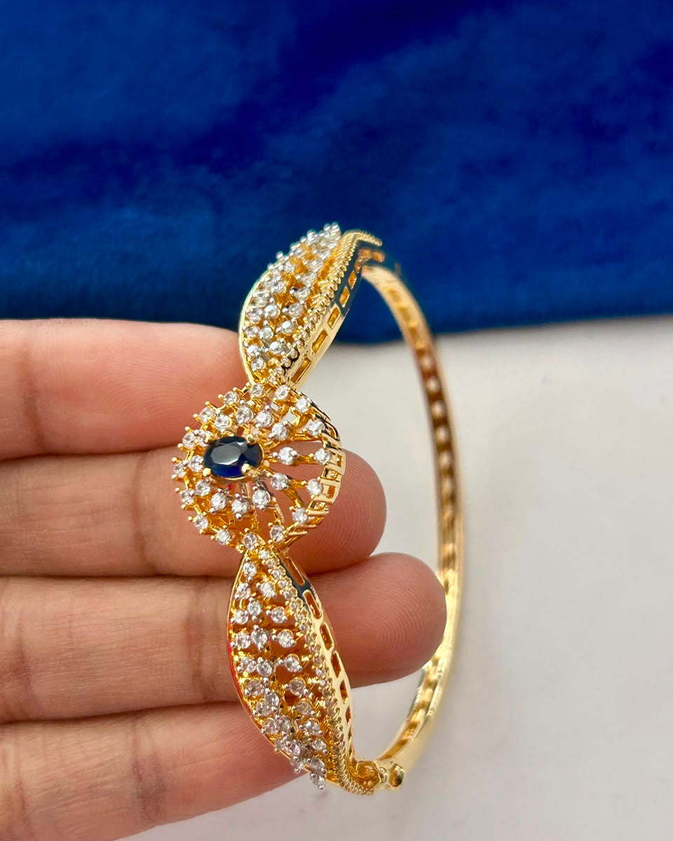 A gold-plated hinged bangle bracelet with pavé-set clear stones and a central dark blue oval gemstone, held in a person's hand against a blue and white background.