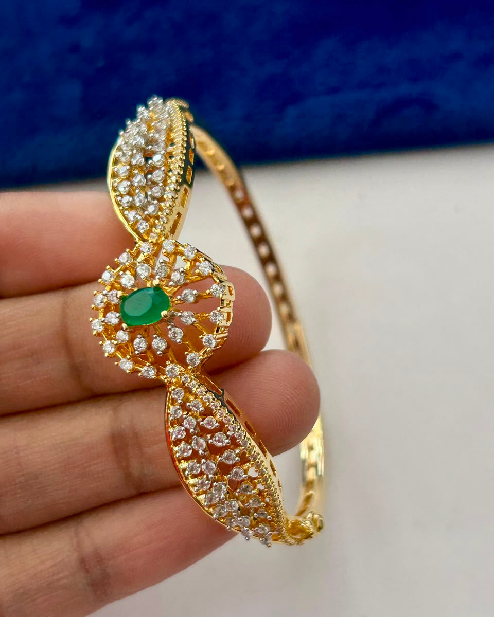 A gold-plated traditional Indian Kundan bangle bracelet with a central green emerald-cut stone and surrounding clear pavé stones, held in a hand against a blue and white background.