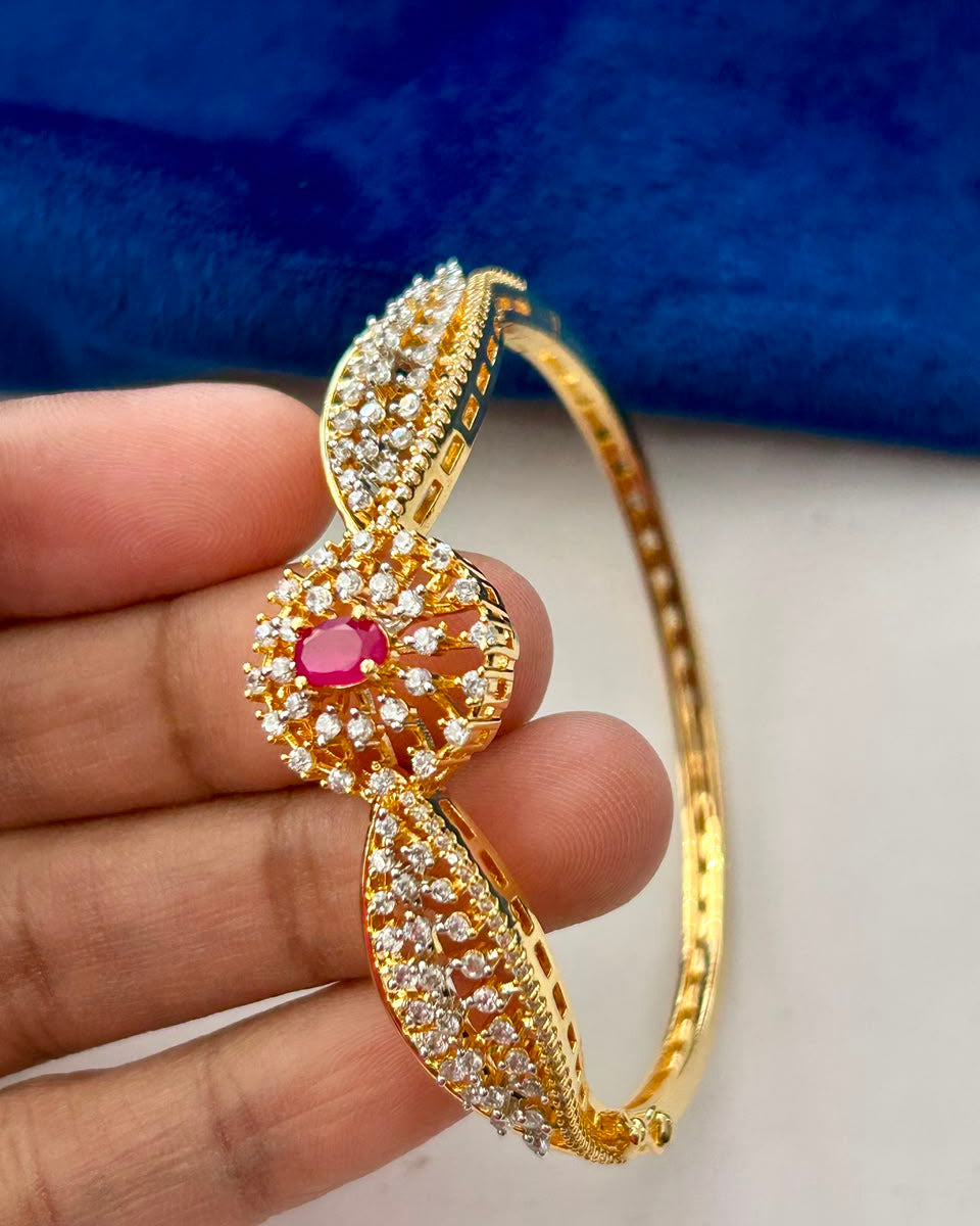 A gold-plated hinged bangle bracelet with pavé-set clear stones and a central pinkish-red oval gemstone, held by a hand against a blue and white background.