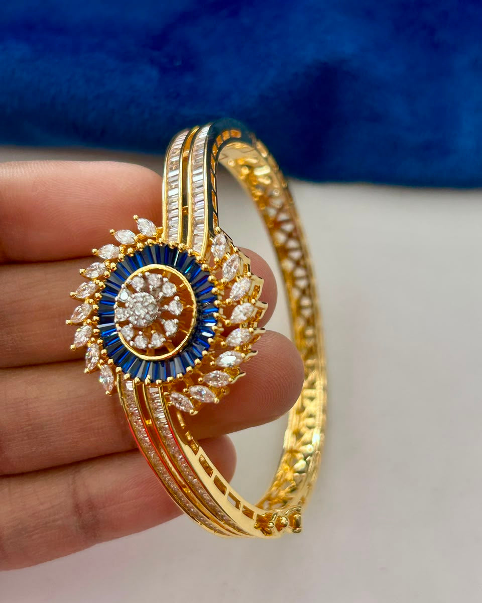 A gold-plated bangle bracelet with a central medallion, held by a hand. The medallion features a blue outer ring, a central diamond cluster, and outer diamond leaves.