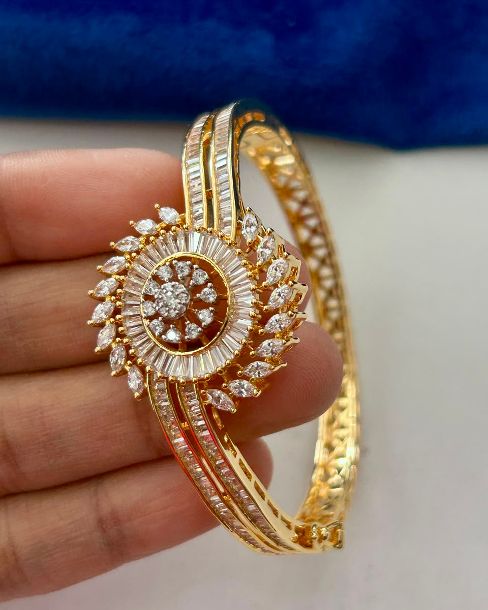 A gold-plated bangle bracelet with intricate diamond and crystal detailing, held in a hand against a white and blue background.