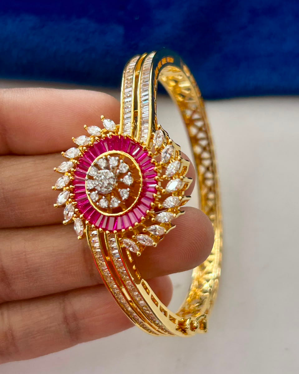 A gold-plated bangle bracelet with a prominent circular pink gemstone centerpiece and clear crystal accents, held by a hand against a blue and white background.