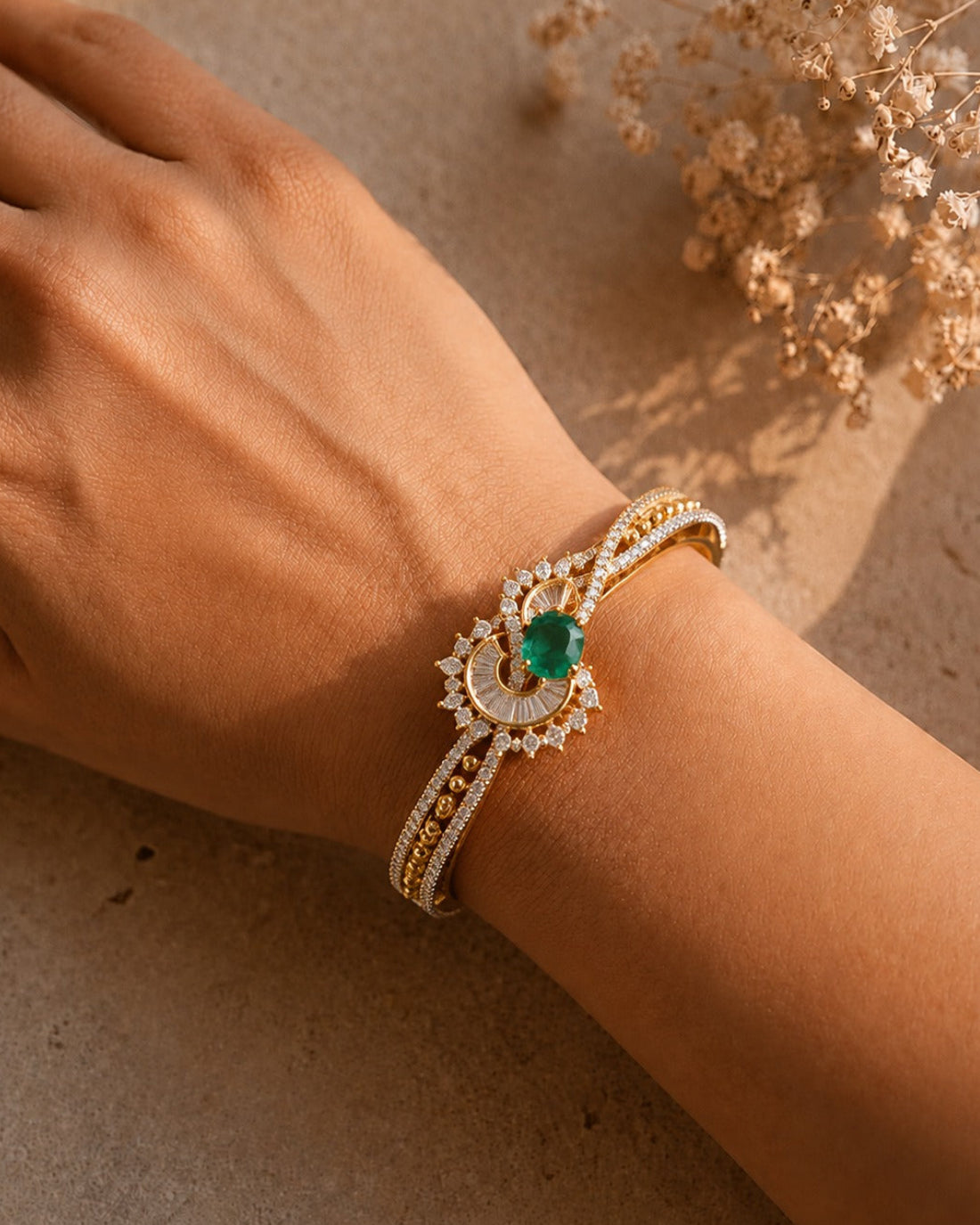 A gold-toned double-band bracelet with a central green emerald and white stones, worn on a person's wrist against a warm beige background with dried flowers.