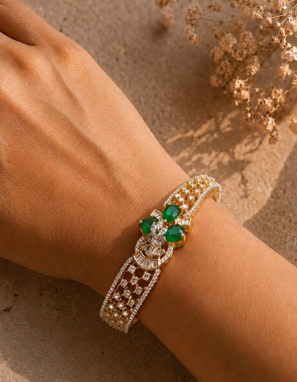 A close-up of a person's wrist wearing an ornate gold-toned cuff bracelet with green emerald-cut stones and pavé-set clear crystals, photographed against a warm beige textured surface with dried flowers in the background.