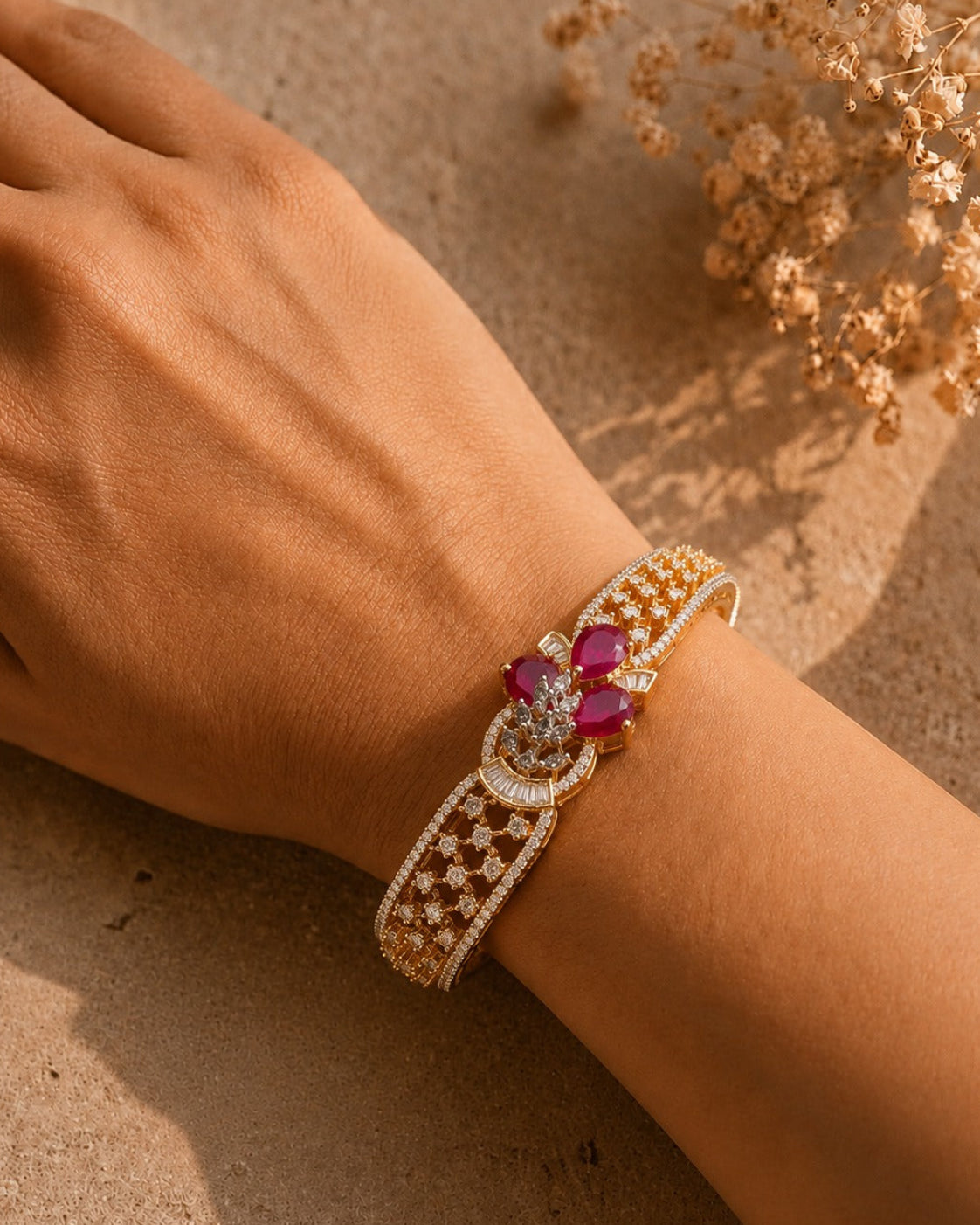A close-up of a person's wrist wearing an ornate gold bracelet with a floral centerpiece featuring deep pink gemstones and clear stones, set against a warm sandy background with dried flowers.