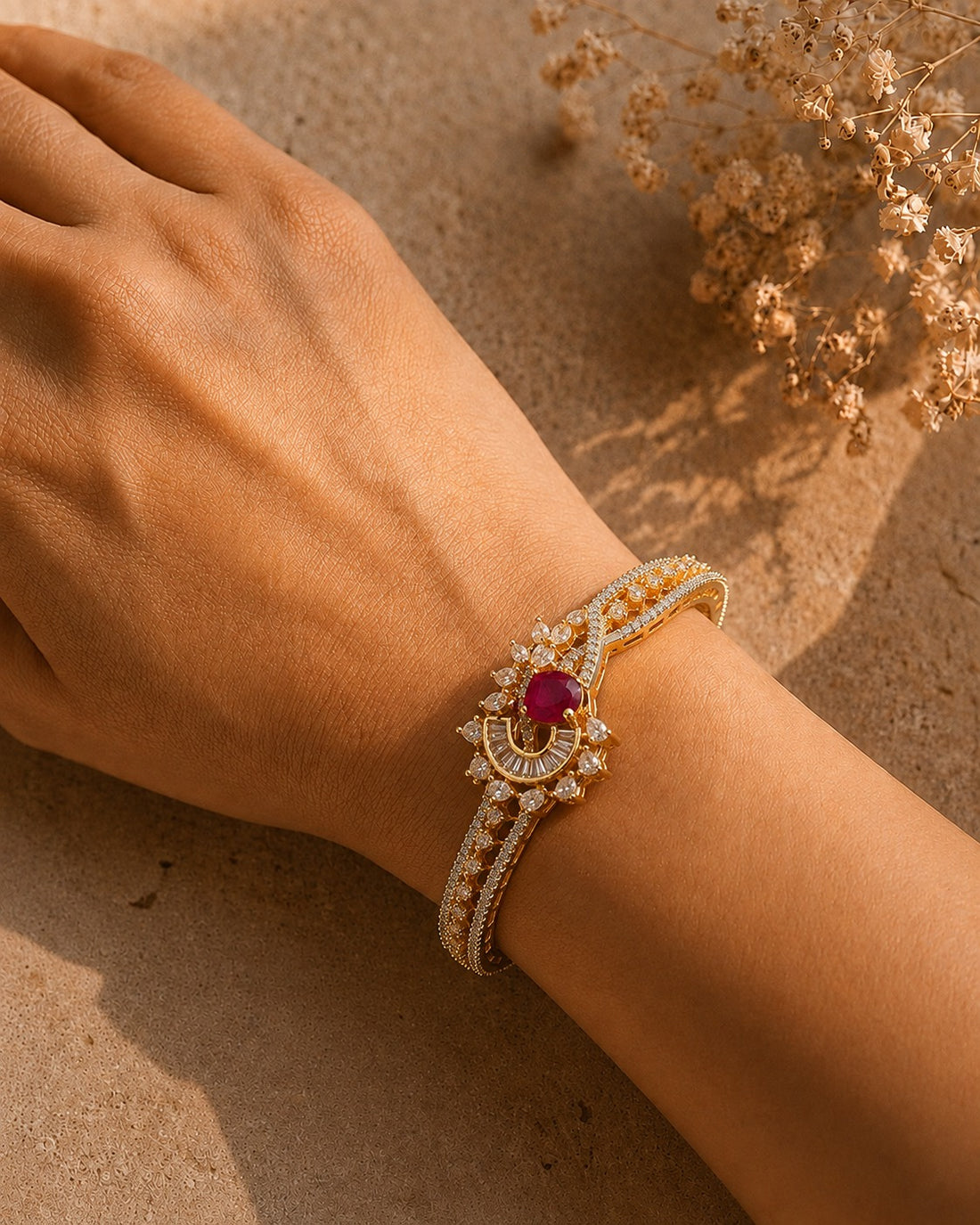 A gold-toned double-strand bracelet with a central round magenta-pink gemstone and surrounding clear stones, worn on a person's wrist against a warm sandy background.