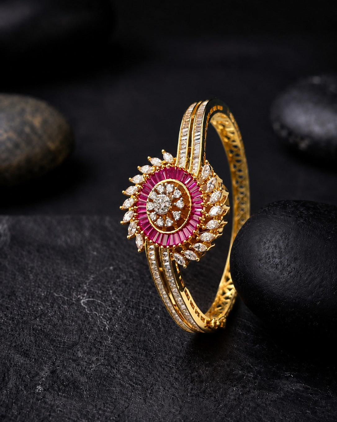 A gold-plated bangle bracelet featuring a circular floral centerpiece with a vibrant pink outer ring, a central clear stone, and rows of small clear stones, displayed on a dark textured surface with black stones.