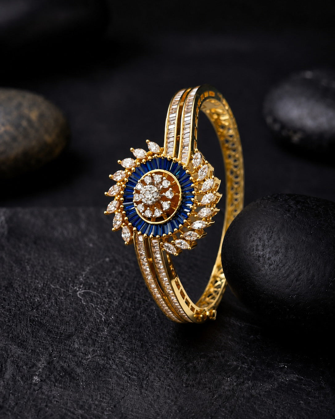 A gold-plated bangle bracelet with a central sunburst design featuring deep blue stones and clear pavé-set stones, displayed on a dark textured surface with smooth black stones.