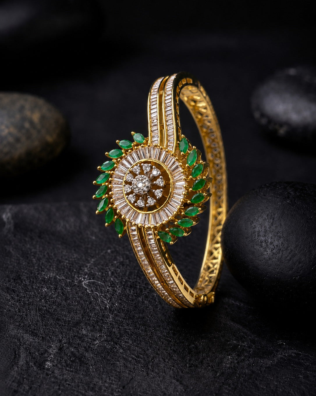 A gold-plated bangle bracelet with a circular floral motif, featuring green emerald-cut stones and a halo of small clear stones, displayed on a dark textured surface with black stones.