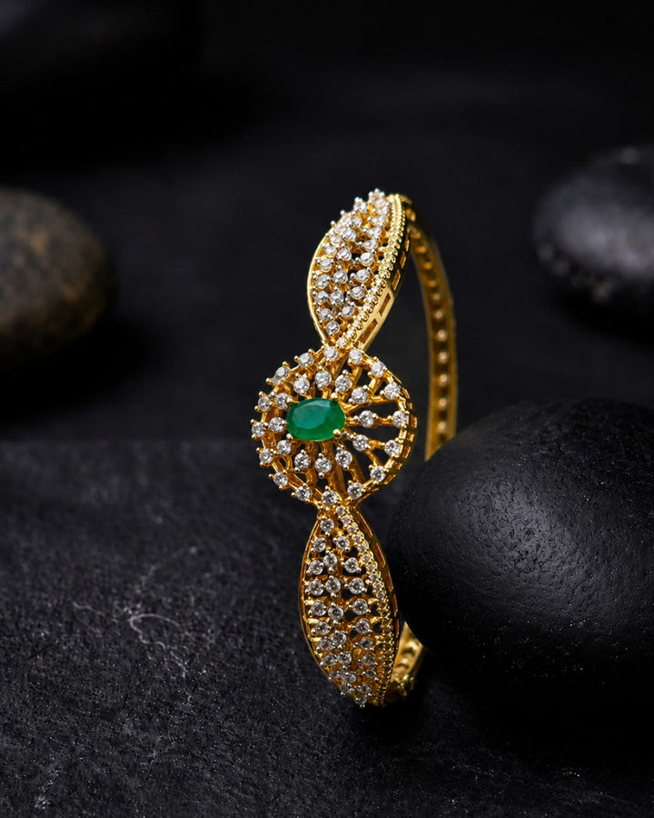 A gold-plated hinged bangle bracelet featuring a central oval cluster of clear pavé stones and a vibrant green oval gemstone, displayed on a dark textured stone surface.