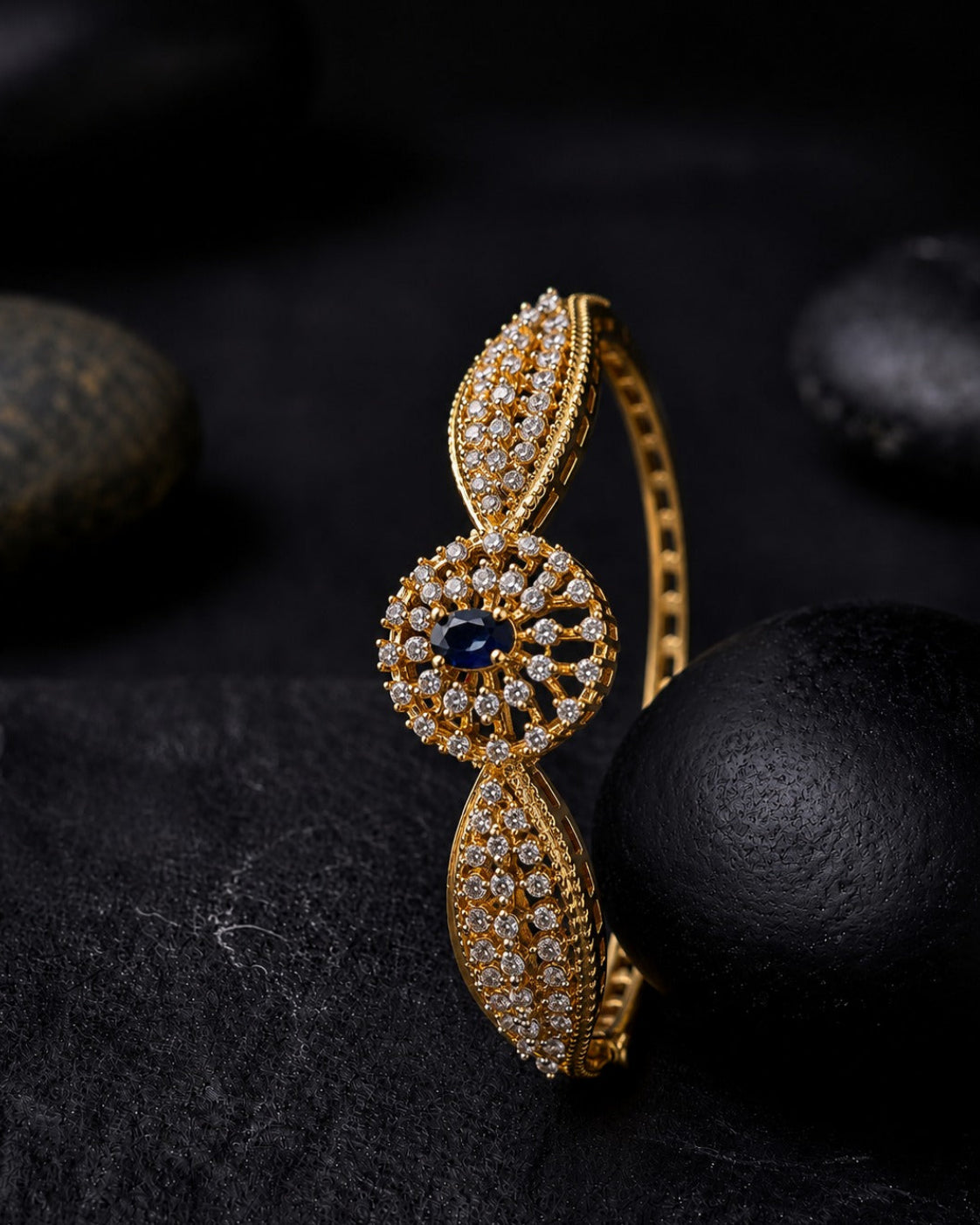 A gold hinged bangle bracelet with pavé-set clear stones and a central dark blue oval gemstone, displayed on a dark textured surface with black stones in the background.