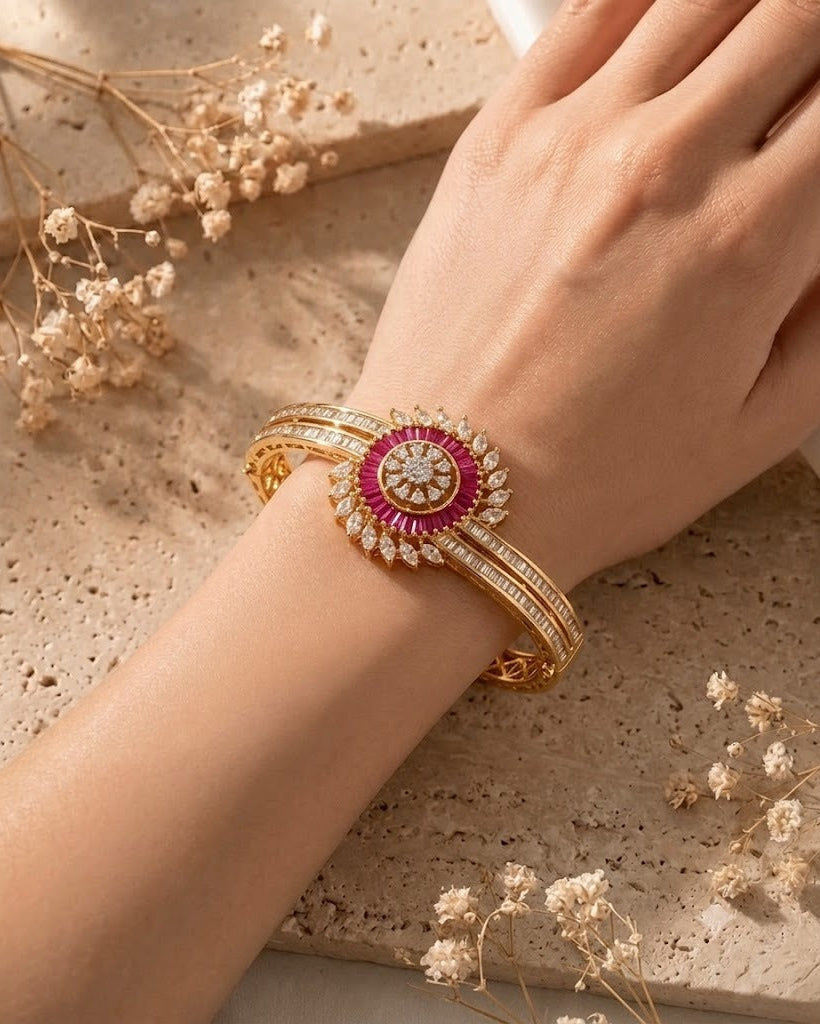 A close-up of a person's hand wearing a gold bangle bracelet with a central floral motif featuring a bright pink center and clear stones, styled on a textured beige stone surface with dried flowers.