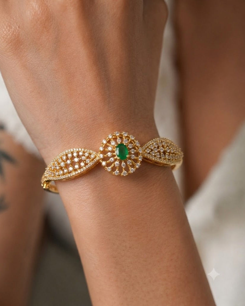 A gold-toned cuff bracelet with a central oval green gemstone and surrounding pavé-set clear stones, shown worn on a person's wrist.