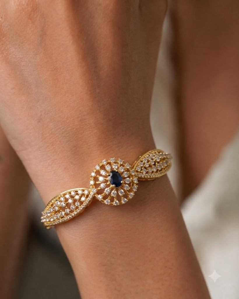 A close-up of a gold-toned cuff bracelet with a central round motif set with clear stones and a dark blue center stone, worn on a person's wrist.