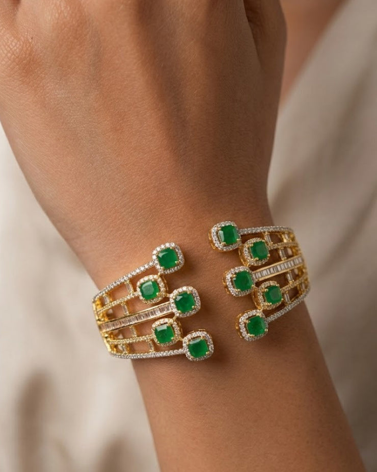 A close-up of a gold cuff bracelet with multiple green emerald-cut stones and small clear accent stones, worn on a person's wrist against a soft beige background.