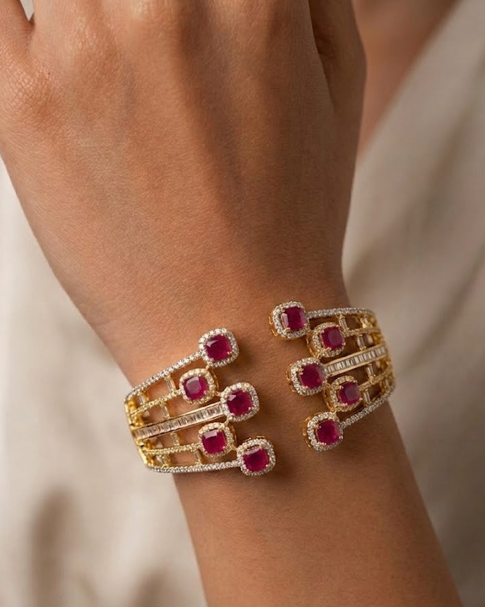 A close-up of a gold cuff bracelet with multiple rows of round-cut pinkish-red gemstones and small clear stones, worn on a person's wrist against a soft beige background.