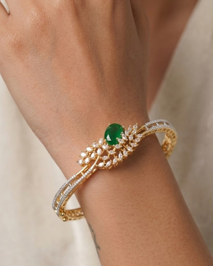 A close-up of a gold bangle bracelet with a central green oval gemstone and rows of small clear stones, worn on a person's wrist against a soft cream fabric background.