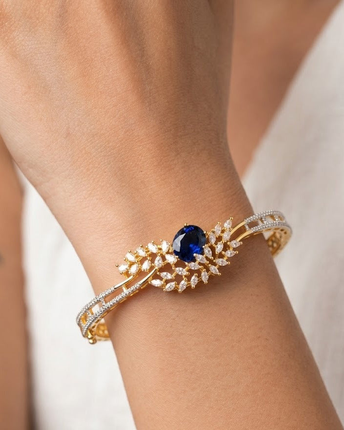 A close-up of a gold leaf-shaped cuff bracelet with a central deep blue oval gemstone and pavé-set clear stones, worn on a person's wrist.