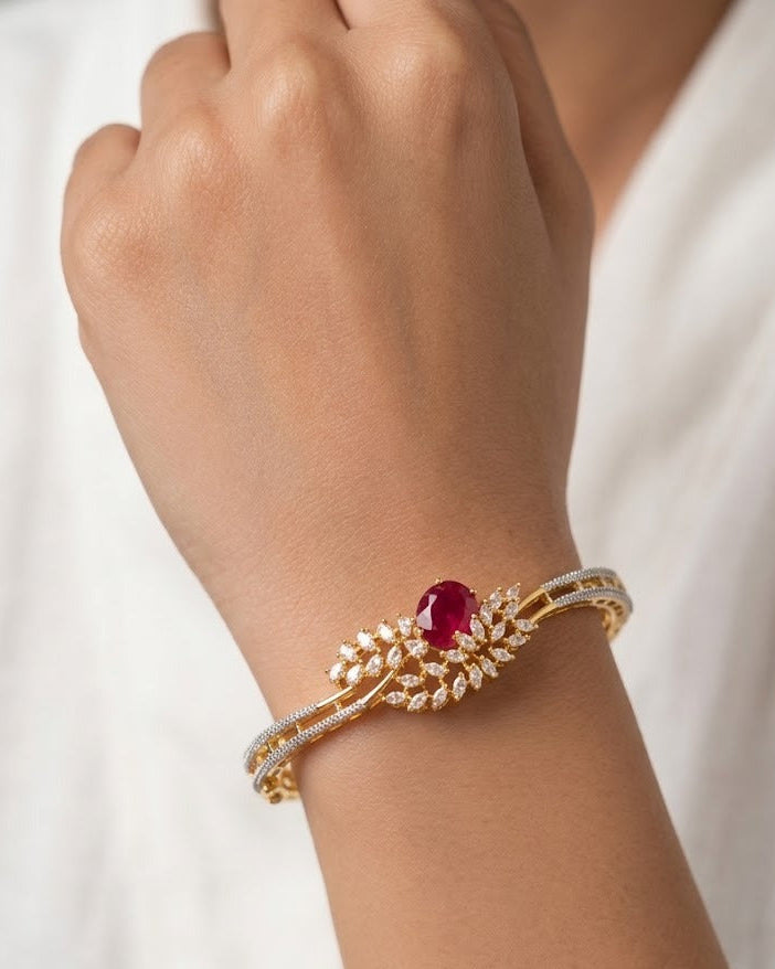 A gold-toned leaf-shaped cuff bracelet with a central oval red gemstone and pavé-set clear stones, shown worn on a person's wrist against a soft white background.