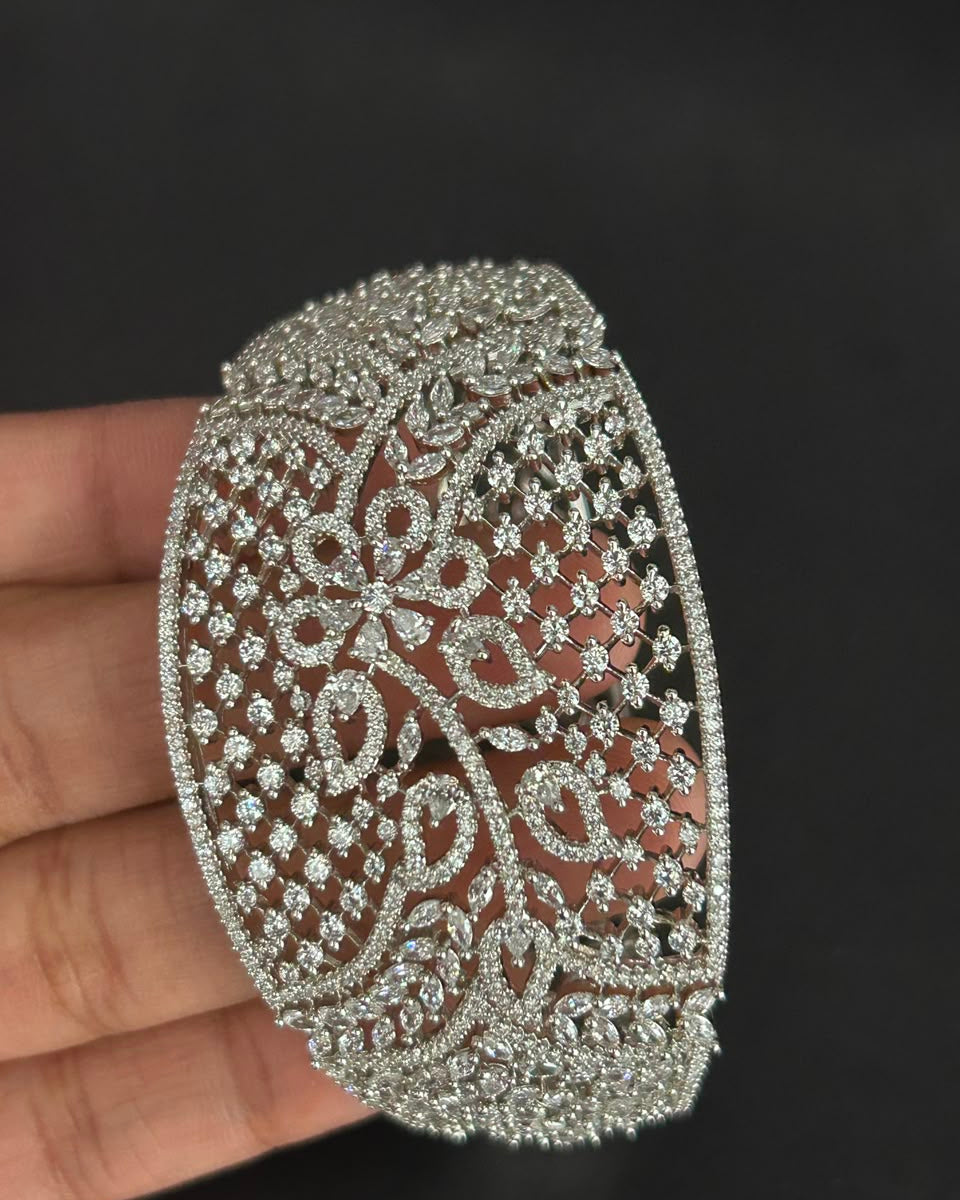 A wide silver-toned filigree bangle bracelet with intricate floral and scroll patterns set with clear stones, held in a hand against a dark background.