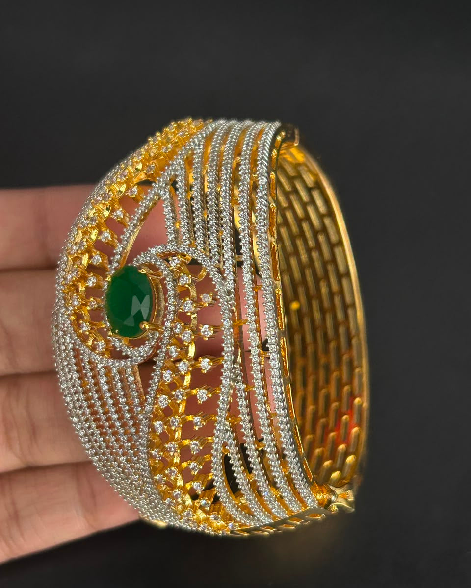 A wide gold-toned bangle bracelet with multiple rows of clear pavé stones and a central green oval gemstone, shown resting on a dark background with a hand holding it for scale.