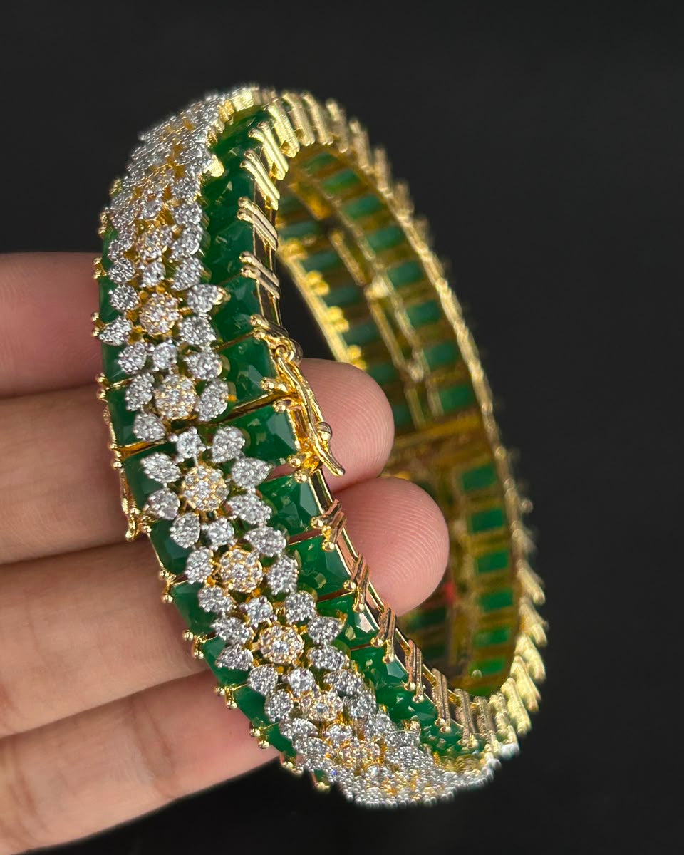 A gold-plated tennis-style bangle bracelet with rows of green emerald-cut stones and clear pavé stones, held in a hand against a black background.