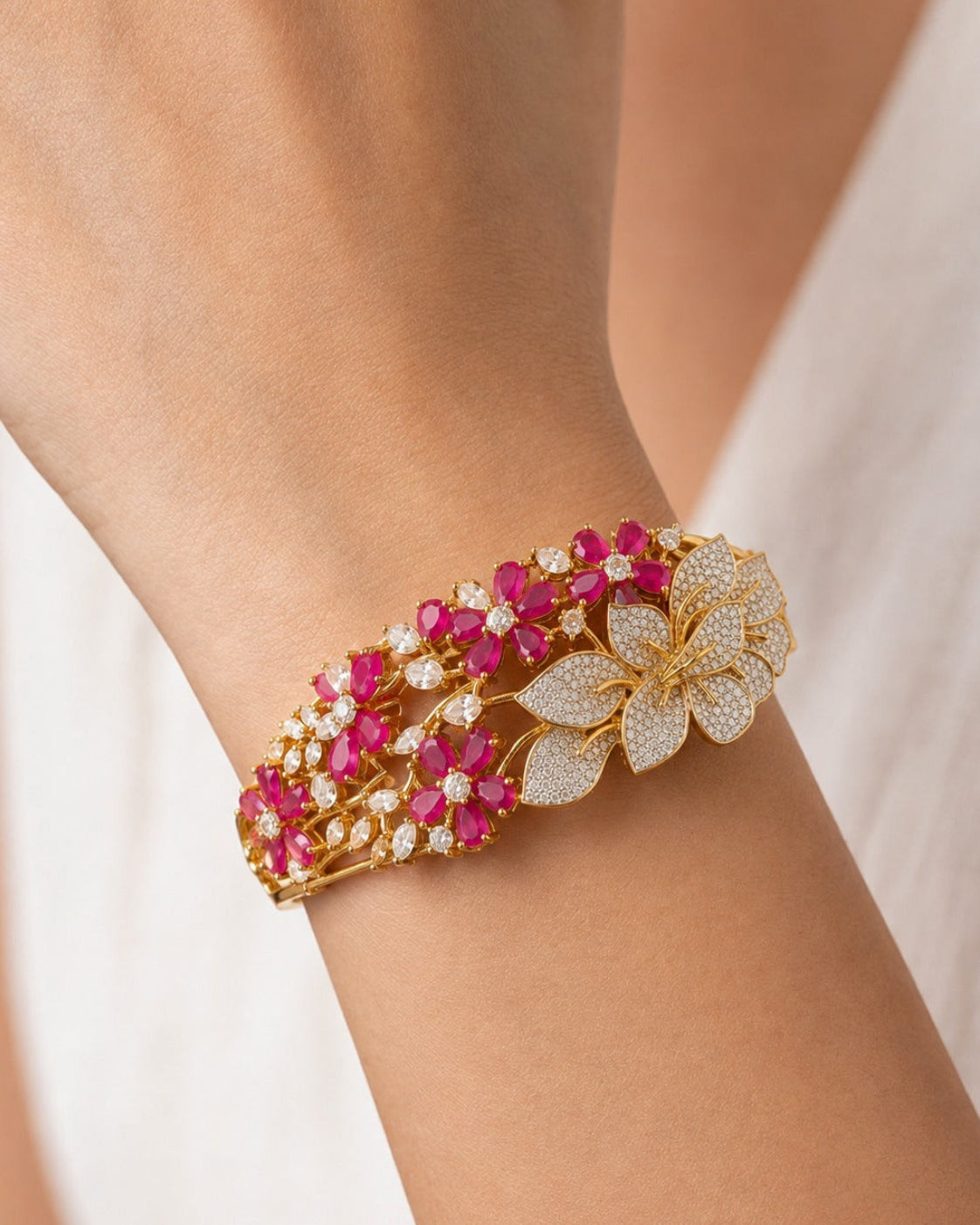 A close-up of a gold-toned floral cuff bracelet with bright pink gemstones and pavé-set clear stones, worn on a person's wrist against a soft white background.