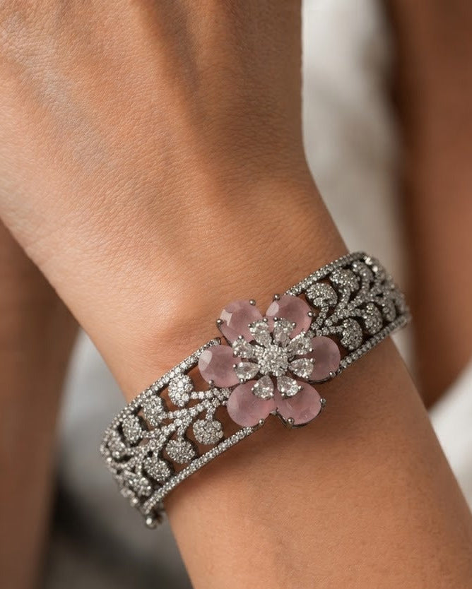 A close-up of a silver-toned wide cuff bracelet with a central pink flower motif and sparkling clear stones, worn on a person's wrist.