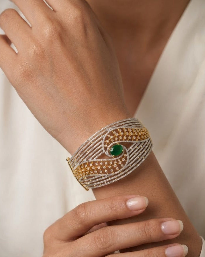 A close-up of a person's wrist wearing a wide cuff bracelet with intricate gold and silver beaded patterns and a central green stone.