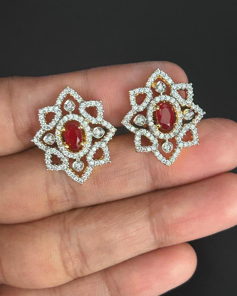 A pair of ornate silver-toned stud earrings with oval red gemstones and surrounding clear stones, displayed on a person's fingers against a dark background.