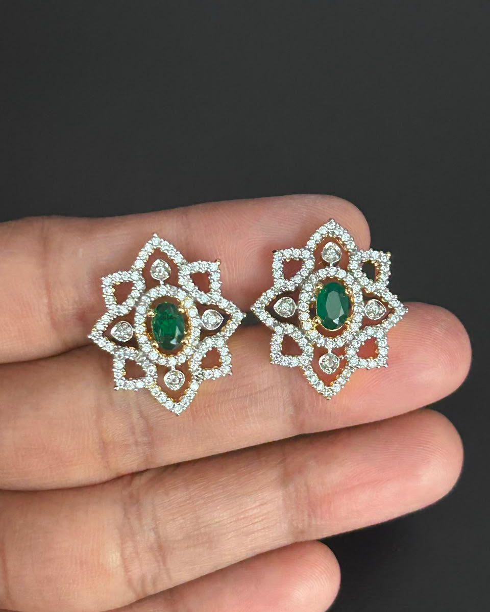 A pair of ornate floral stud earrings with oval green emerald-cut center stones and surrounding pavé-set clear stones in a gold-tone setting, shown resting on a person's fingers against a dark background.