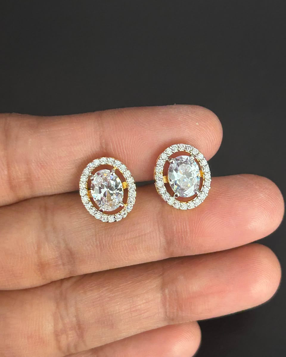 A pair of oval stud earrings with clear gemstones in a yellow-gold setting, held between fingers against a dark gray background.