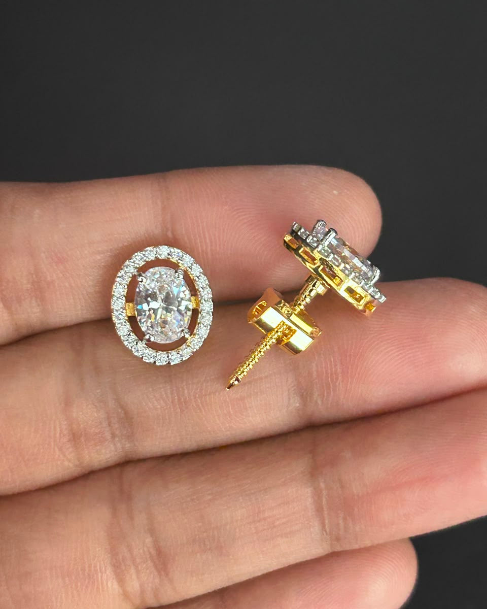 A pair of oval halo stud earrings with clear gemstones, one shown from the front and the other from the side/back, resting on a person's fingers against a dark background.